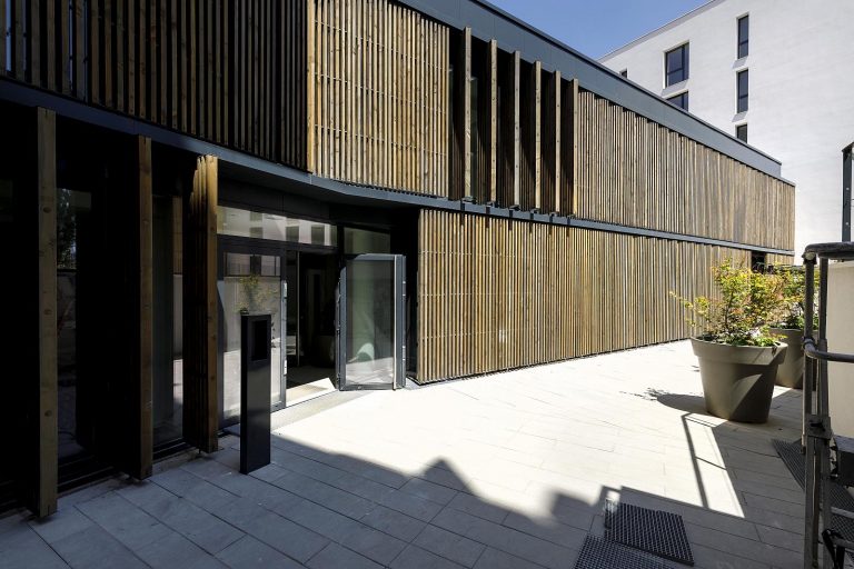 Vertical timber sunbreakers along a building facade, several panels folded open onto a paved terrace
