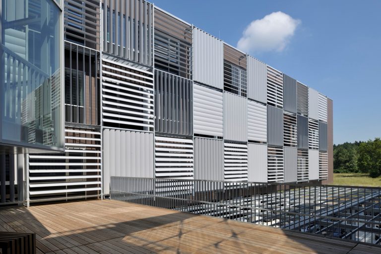 Building facade clad in movable white and bronze aluminium shutter panels set at varying angles, seen from a wooden roof terrace