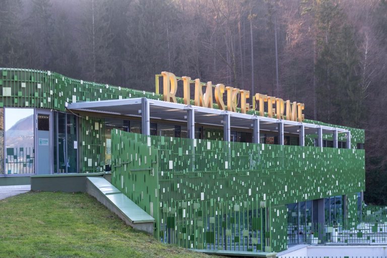 Retractable canopy over the terrace of a spa building with a green mosaic facade, with a wooded hillside behind