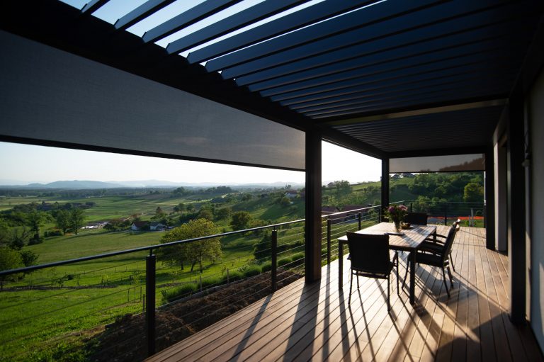 View from under a bioclimatic pergola with side screen blinds, across a wooden terrace to rolling green countryside