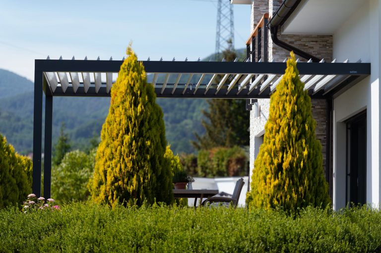 Free-standing bioclimatic pergola with open louvres beside a house, framed by conifers with hills behind