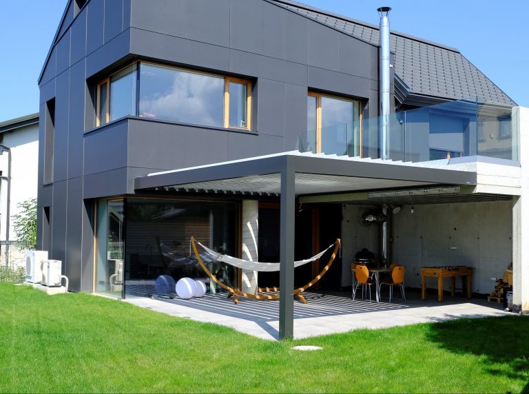 Bioclimatic pergola attached to a dark-clad modern house, sheltering a terrace with a hammock and a table football