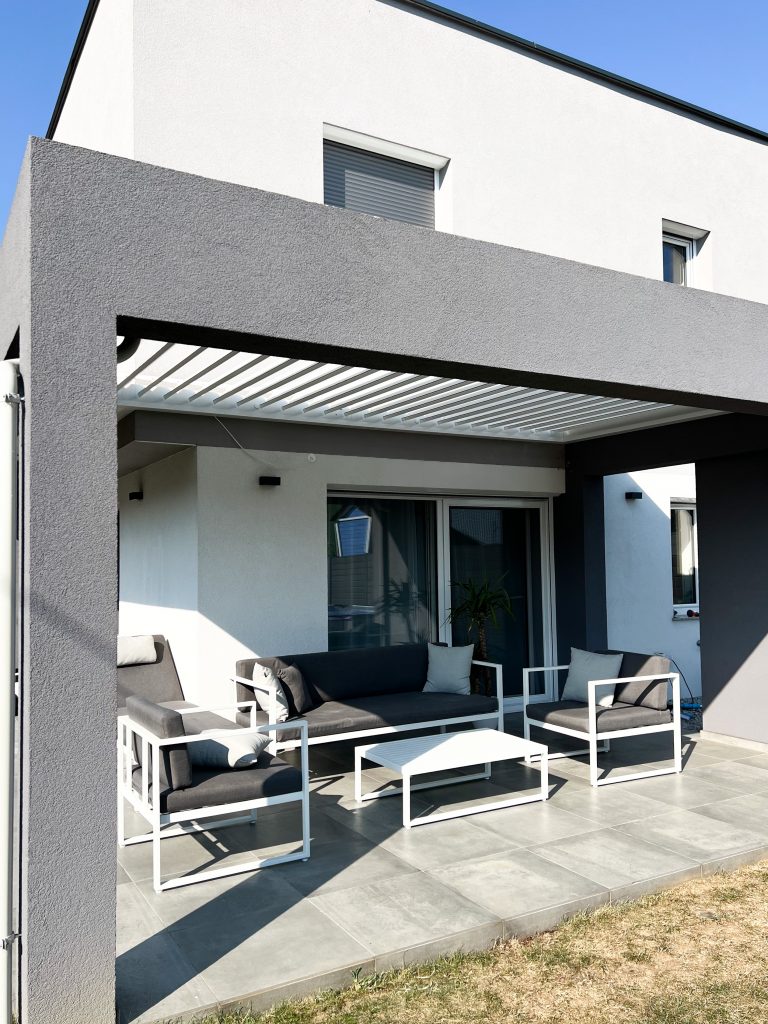White bioclimatic pergola with open louvres over a paved terrace with white lounge furniture, beside a modern house