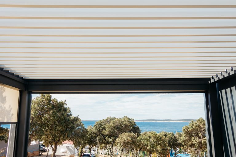 Cream bioclimatic pergola louvres above a terrace opening onto pine trees and the sea