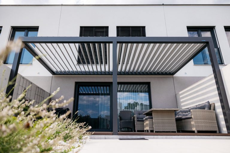 Bioclimatic pergola with open louvres over a terrace with rattan lounge furniture, seen from the garden