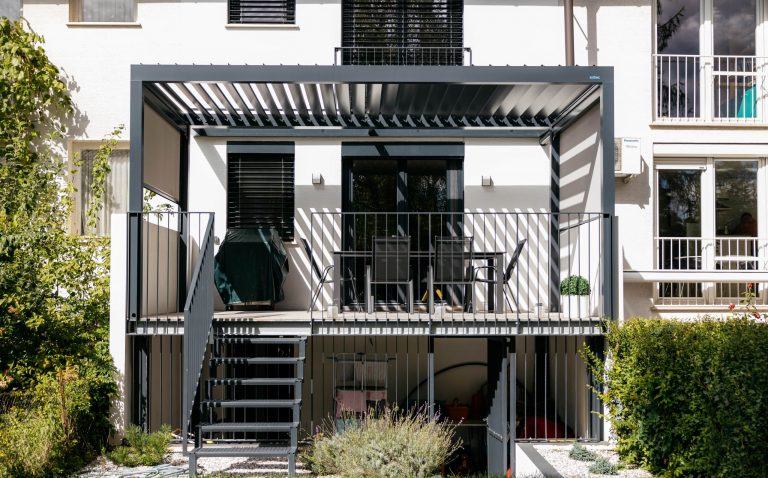 Bioclimatic pergola with open louvres over the first-floor balcony of a white house, with side screen blinds and a steel staircase