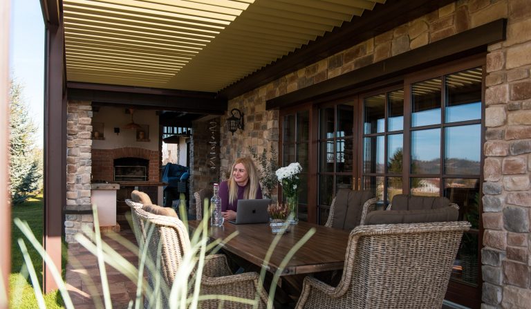 Bioclimatic pergola over a stone-clad terrace with a wood-fired oven and a dining table, with someone working at a laptop