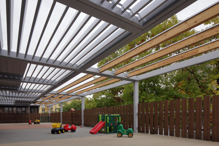 Large aluminium and timber canopy shading a nursery playground, with ride-on toys and a slide beneath the open roof