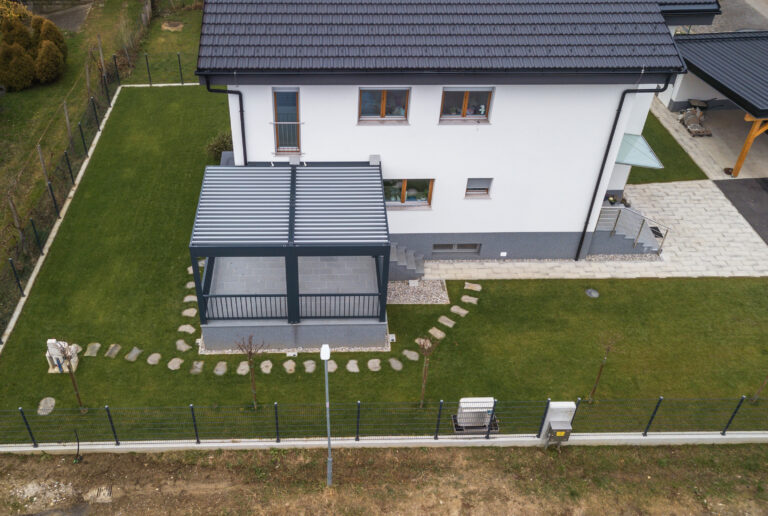 Aerial view of a detached white house with a dark tiled roof and an anthracite bioclimatic pergola with closed louvres over the terrace