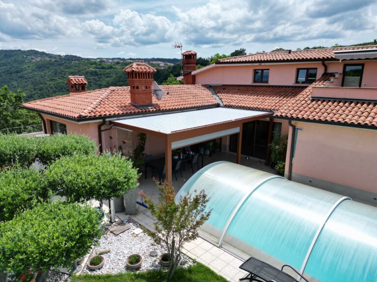 Bioclimatic pergola over the terrace of a hillside house with a tiled roof, beside a covered swimming pool