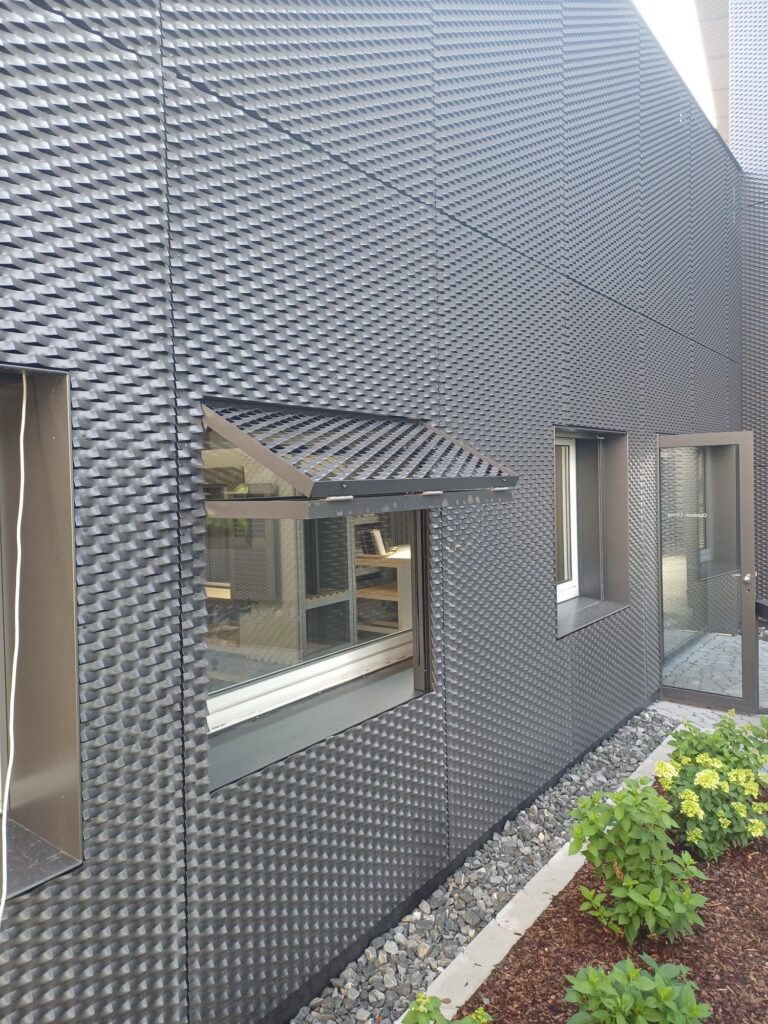 Dynamic facade of dark textured aluminium panels, with one panel lifted open above a window